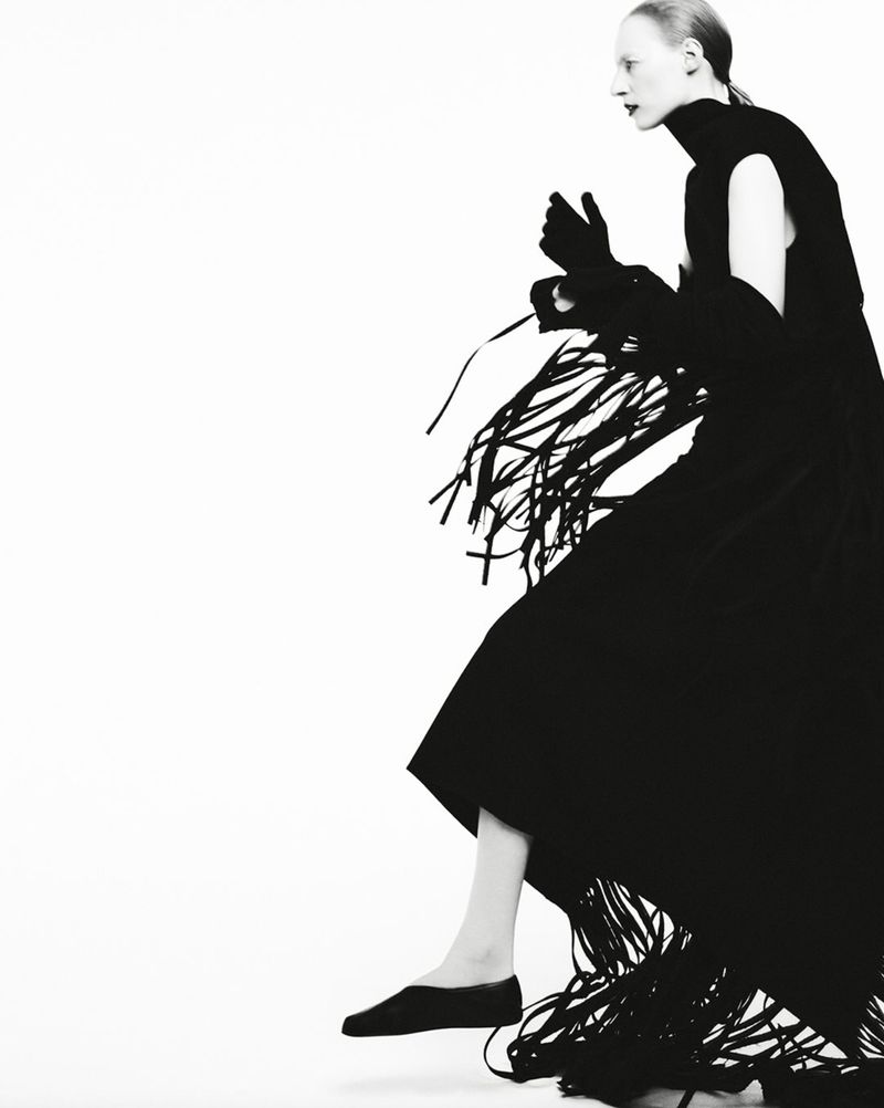 A slender fashion model poses in a black turtleneck dress and long fringed shawl or overskirt against a white backdrop in a studio setting.