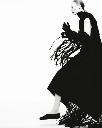 A slender fashion model poses in a black turtleneck dress and long fringed shawl or overskirt against a white backdrop in a studio setting.
