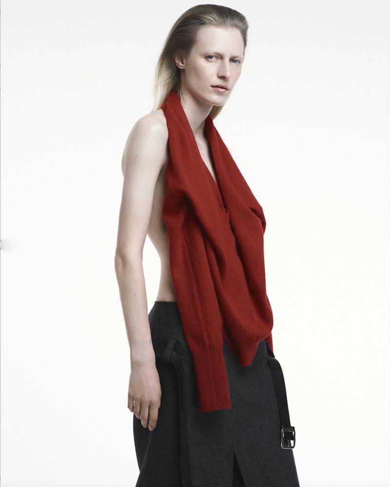 Fashion portrait of a blonde woman in a red knit top and charcoal skirt posing in a photo studio with a white background.