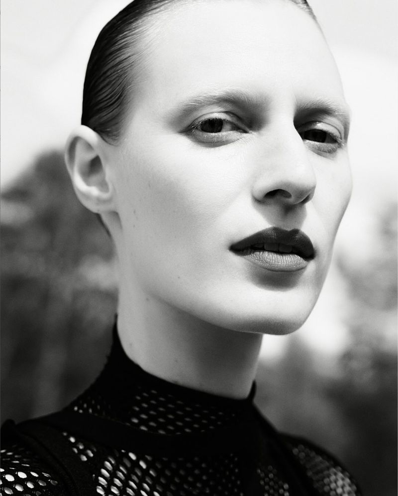Monochromatic portrait of a fair-skinned adult female with slicked-back hair wearing a dark top, with a blurry abstract background.