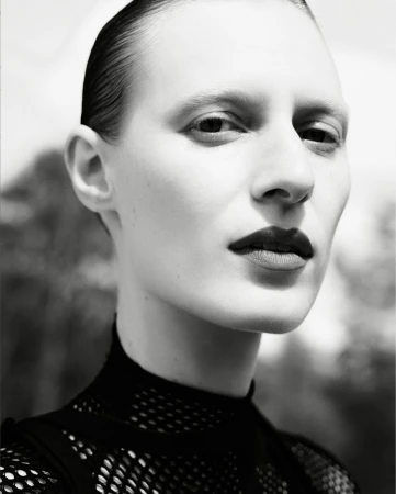 Monochromatic portrait of a fair-skinned adult female with slicked-back hair wearing a dark top, with a blurry abstract background.
