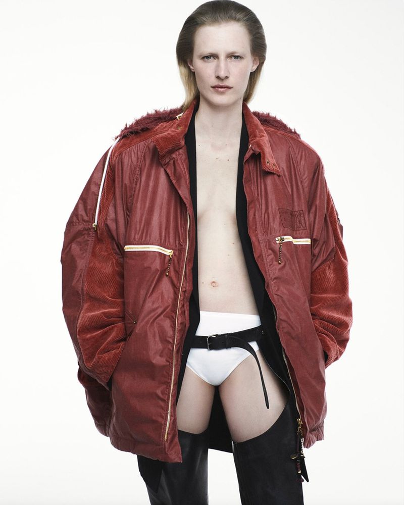 Fashion shot of a model posing in a red jacket, black top, white underwear, and black chaps in front of a white studio backdrop.