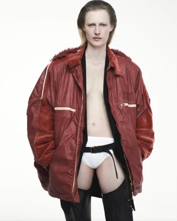 Fashion shot of a model posing in a red jacket, black top, white underwear, and black chaps in front of a white studio backdrop.