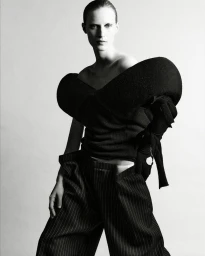 Model Julia Nobis posing in a sculptural off-the-shoulder black top and wide pinstripe trousers against a white seamless studio backdrop.