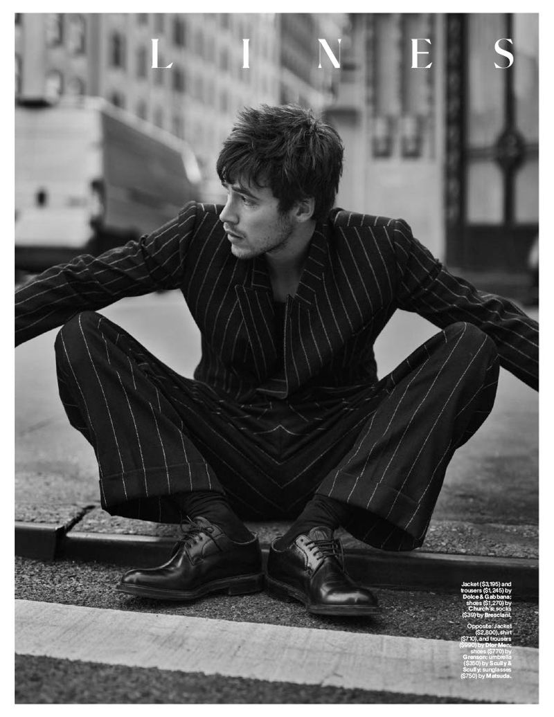 Black and white portrait of a man in a pinstripe suit, seated outdoors on a city street with buildings and a truck in the background.