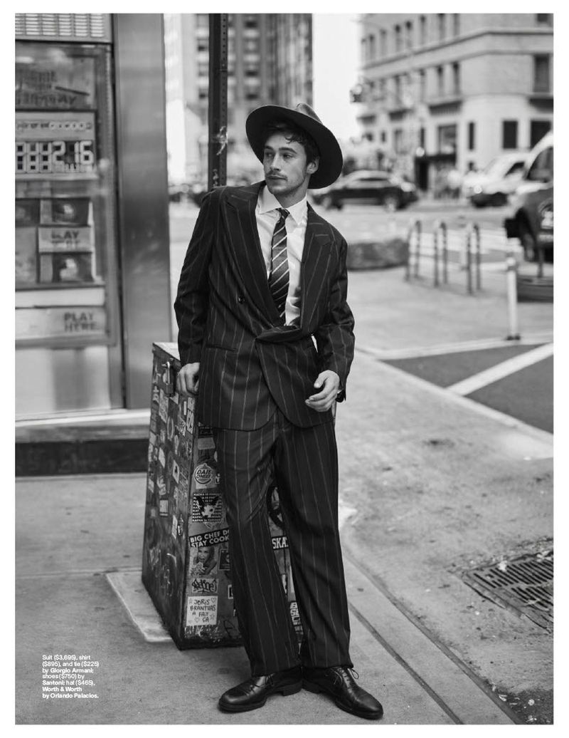 Black and white shot of a stylish man in a pinstripe suit and hat leaning against a vintage-style payphone on a city street.