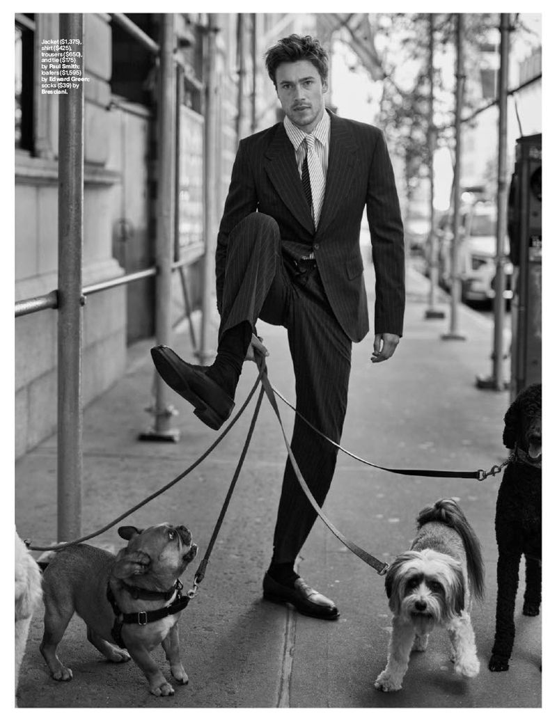 A man in a suit stands on a city sidewalk with multiple dogs on leashes, with a light-toned building and street visible in the background.