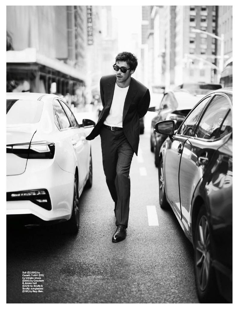 Monochrome shot of a man in a suit walking down a city street between rows of parked cars in bright daylight.