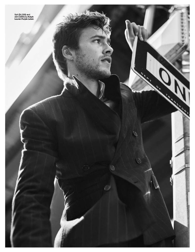 Black and white portrait of a young adult male wearing a blazer, holding a one-way sign outdoors on a city street with sunlight.