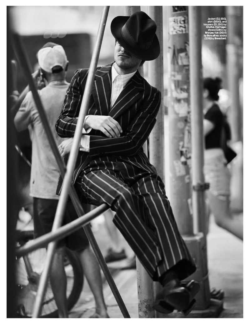 Monochrome portrait of a stylish adult man wearing a striped suit and fedora while resting on scaffolding in an urban environment.