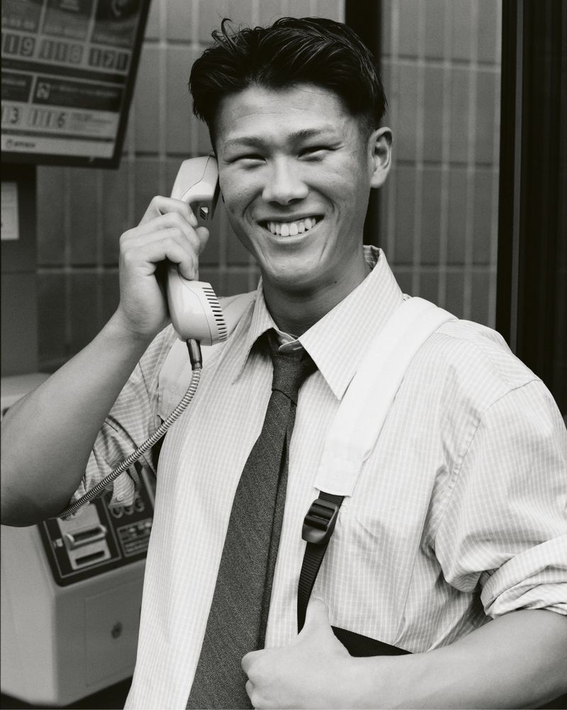 Smiling man in a shirt and tie uses a public phone booth with a telephone receiver in hand indoors in a black and white photograph.