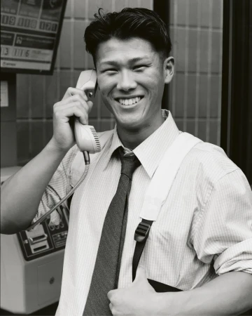 Smiling man in a shirt and tie uses a public phone booth with a telephone receiver in hand indoors in a black and white photograph.