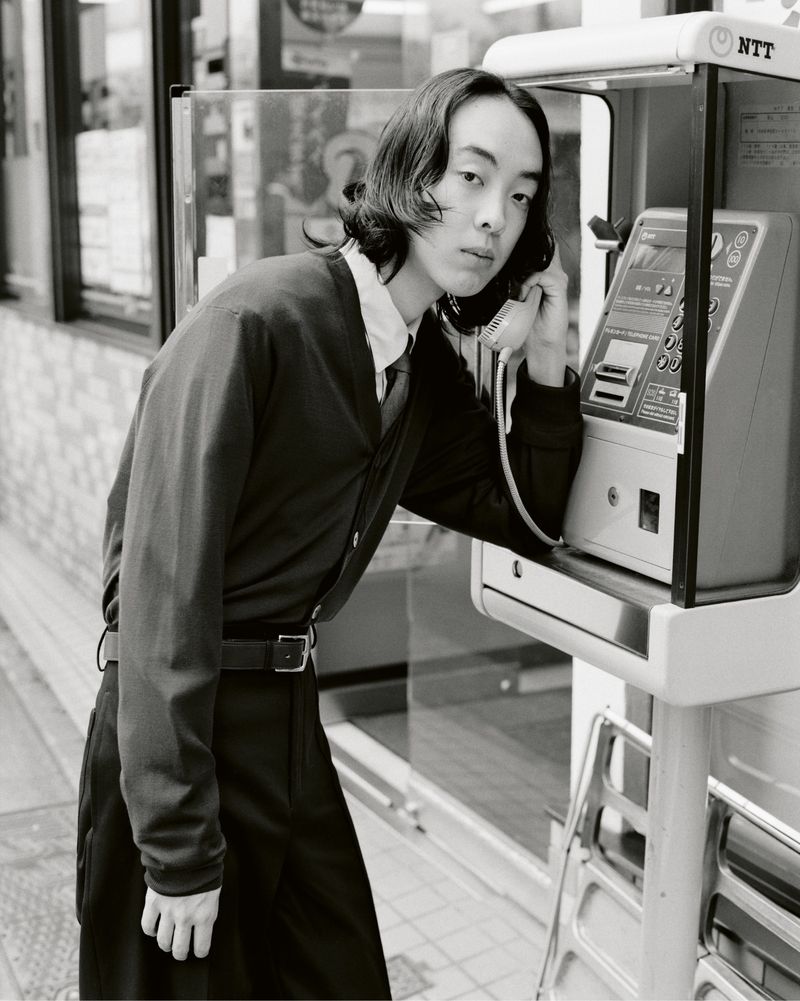 Monochrome portrait of a slender adult man in dark cardigan using a public payphone, set against a neutral city background.