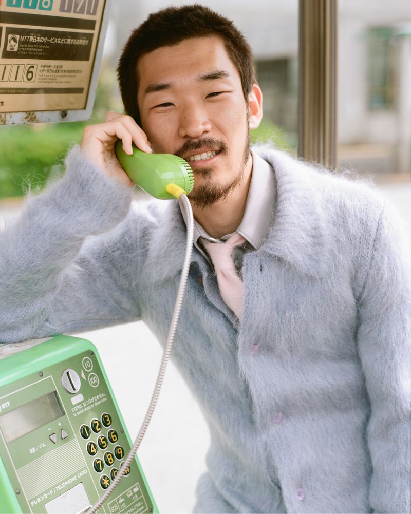 Asian adult male in a fuzzy coat using a green public telephone outside, with natural lighting and a medium shot, creating an engaging portrait.
