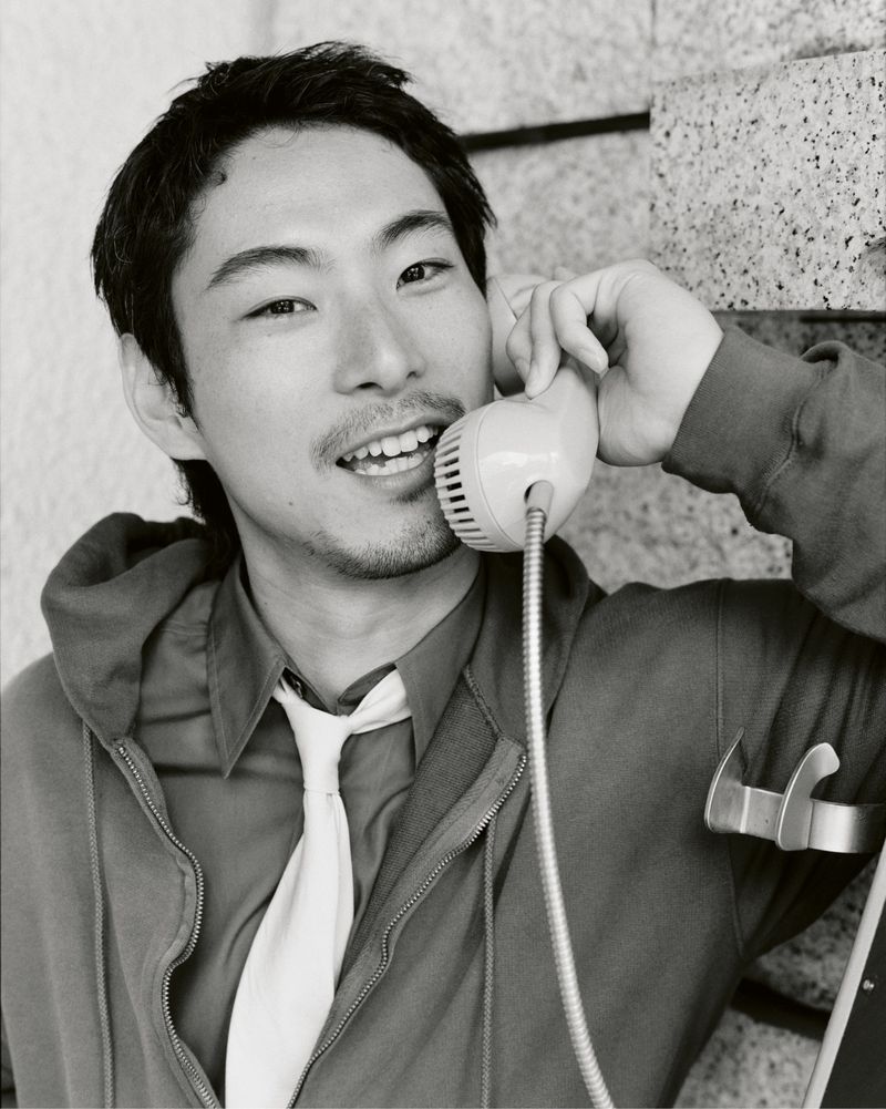 Monochrome portrait of a young man with dark hair talking on a vintage telephone in a gritty urban setting, wearing a hoodie and tie.
