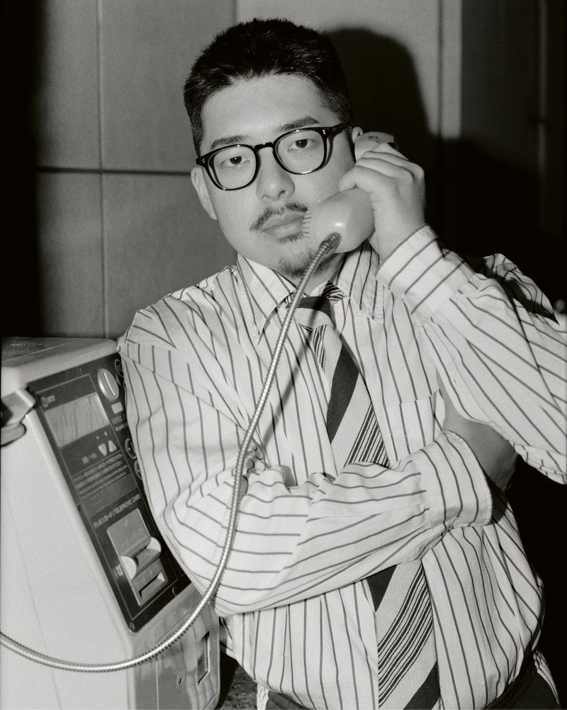 Black and white portrait of a man in a striped shirt using a public payphone, indoors.