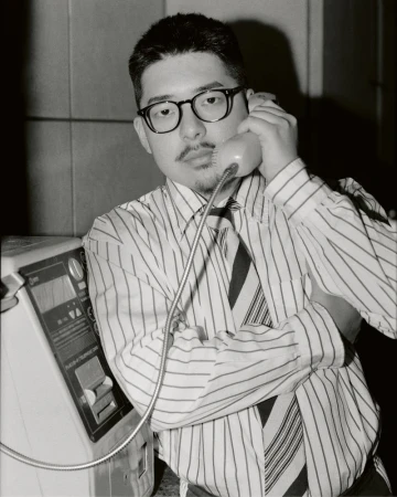 Black and white portrait of a man in a striped shirt using a public payphone, indoors.