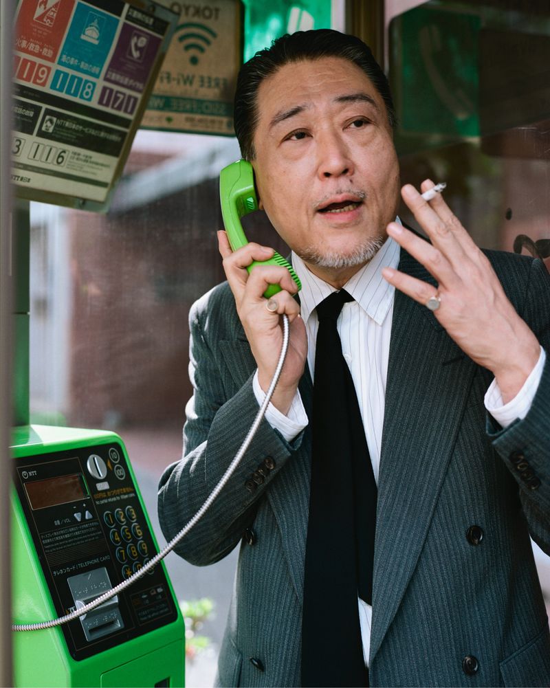 Adult male in business suit inside a phone booth using a green retro phone, smoking, in an urban environment, medium shot.