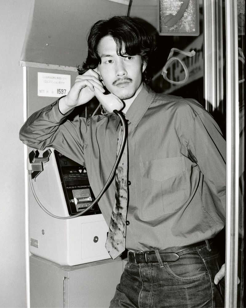 Black and white shot of a man in a red shirt and tie using a public phone booth. Portrait style with a medium shot.