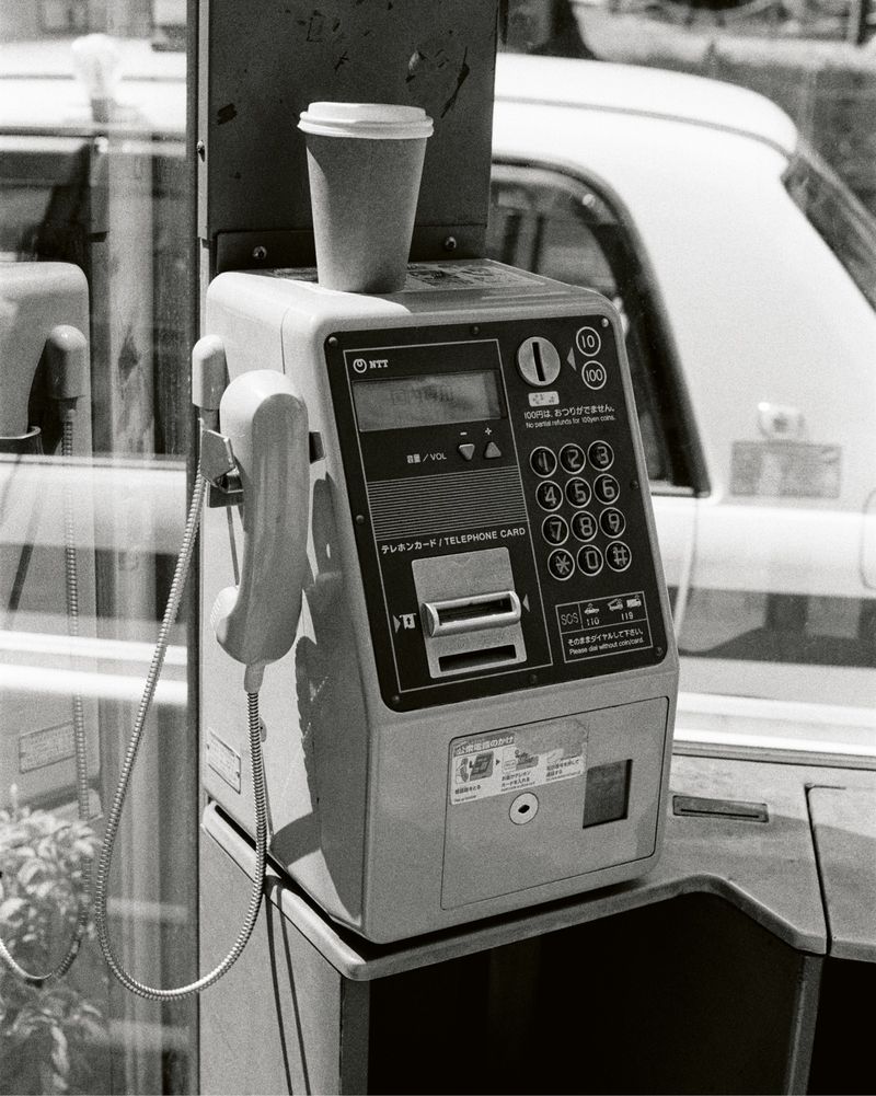 Monochrome image of a public phone booth with a disposable coffee cup on top of the phone, reflecting an outdoor scene.