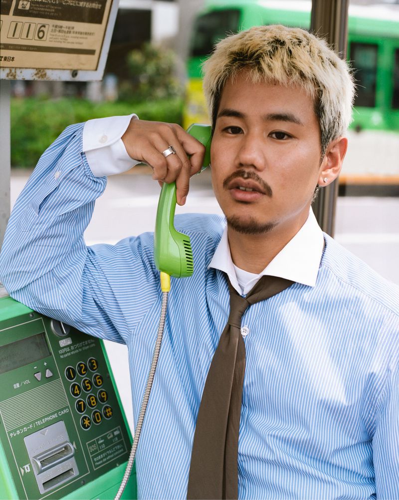 Adult male with dyed-blond hair, wearing a blue striped shirt and brown tie, holds a green payphone handset in an outdoor urban environment.