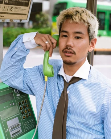 Adult male with dyed-blond hair, wearing a blue striped shirt and brown tie, holds a green payphone handset in an outdoor urban environment.