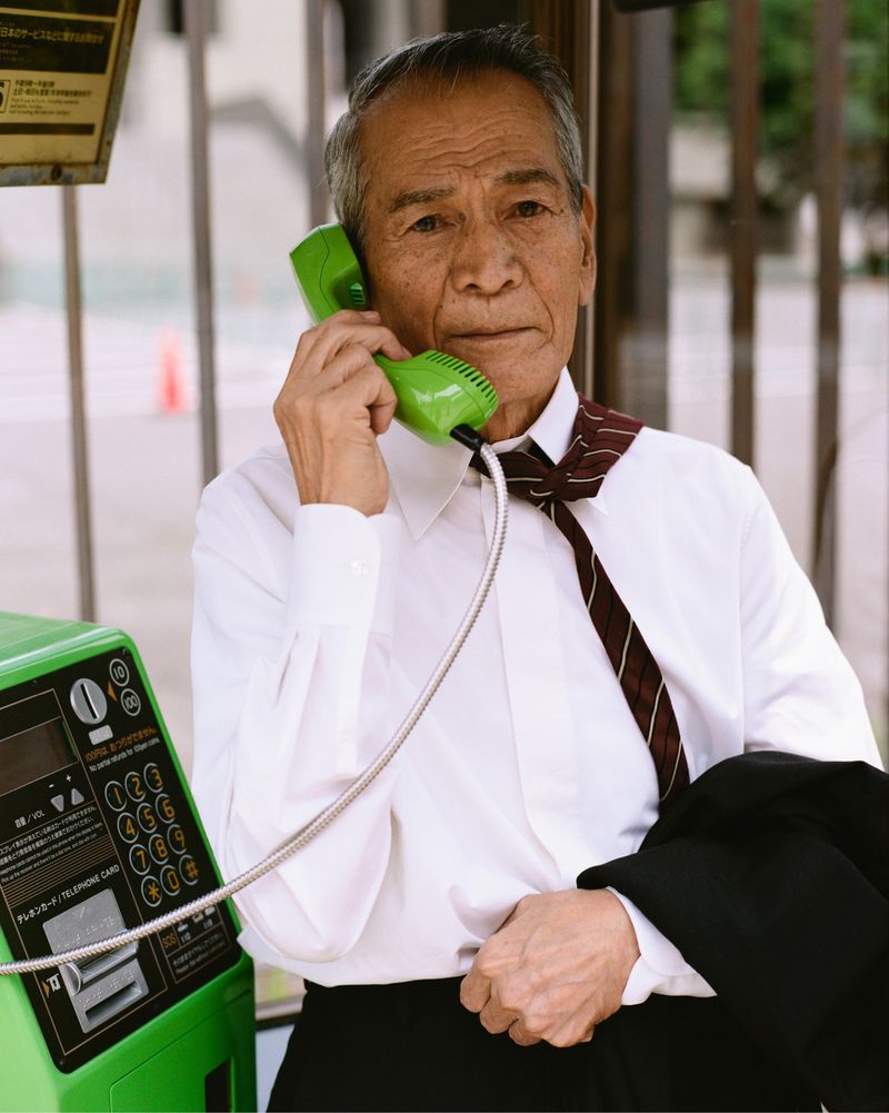 An older Asian man uses a green public telephone, wearing a white shirt and tie, standing in an urban setting on a sunny day.