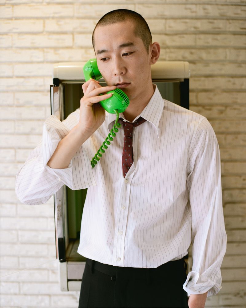 Young man in striped shirt and tie stands inside an indoor public telephone booth, holding a green retro telephone receiver up to his ear.