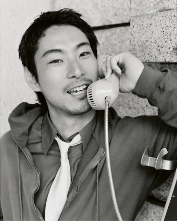 East Asian man smiling while speaking into a corded telephone handset, dressed in a zip hoodie, collared shirt, and white necktie.