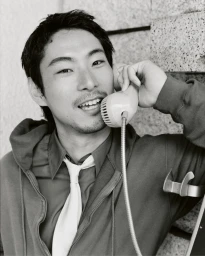 East Asian man smiling while speaking into a corded telephone handset, dressed in a zip hoodie, collared shirt, and white necktie.