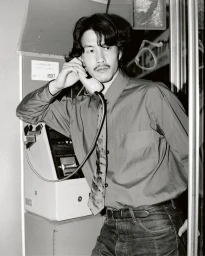 Man with mustache in a button-down shirt using a public payphone inside a booth