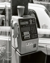 Black and white close-up of a Japanese public payphone inside a booth with a paper takeaway coffee cup resting on top.