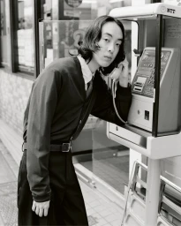 Man with dark medium-length hair wearing a dark cardigan and trousers using a public payphone on a street.