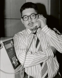 Man wearing a striped shirt and glasses holding a public payphone handset to his ear.