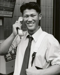 Smiling man in striped shirt and tie holding a public payphone receiver outdoors