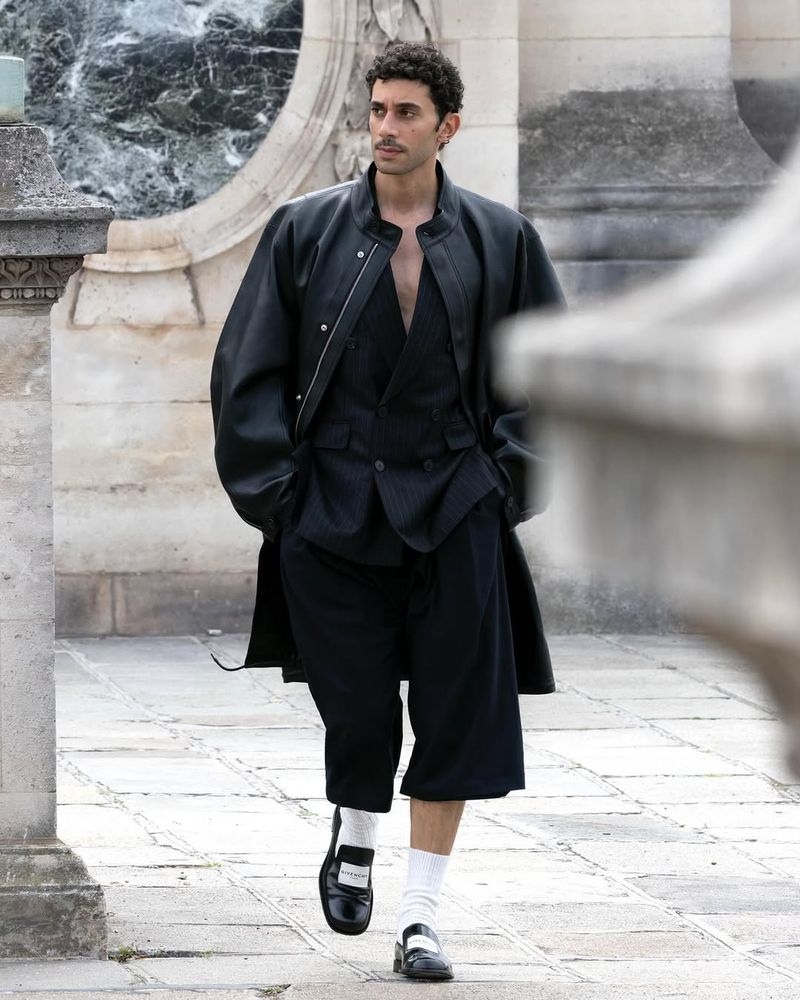 A fashionable man in a black jacket, vest, and shorts walks outdoors, showcasing a contemporary high fashion look with neutral tones.