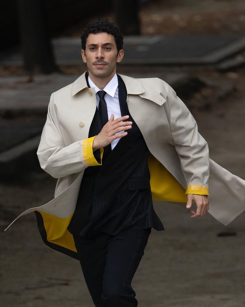 Adult male running, wearing a beige trench coat over a black suit with white shirt and black tie in an outdoor setting with blurred background.