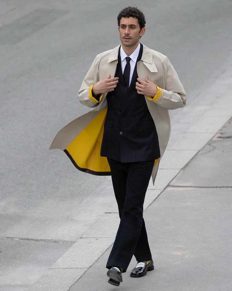 Man in beige trench coat, black suit, and black loafers strides on a concrete surface, embodying modern fashion and urban style.