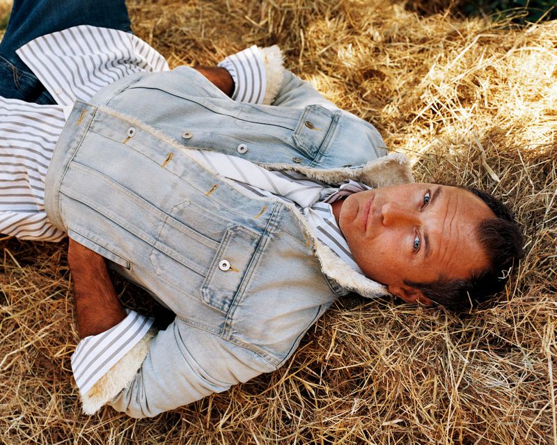Adult male laying in dry grass wearing a denim jacket and striped button-down shirt. Natural outdoor setting, eye-level angle.