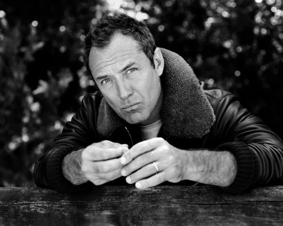 Black and white portrait of adult male with short hair, wearing a leather jacket, leaning against wooden surface in an outdoor setting.