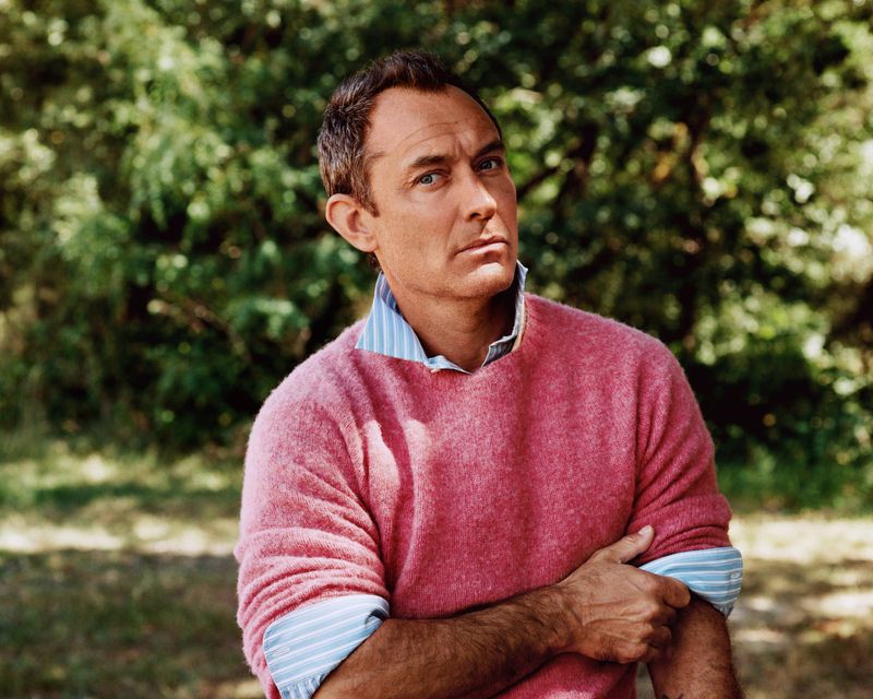 Jude Law wearing a pink wool crewneck sweater over a striped blue shirt outdoors against a leafy green woodland background