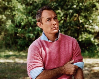 Jude Law wearing a pink wool crewneck sweater over a striped blue shirt outdoors against a leafy green woodland background