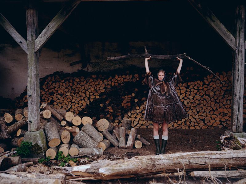 Fashionable young woman in a brown coat stands outdoors in front of firewood, holding a branch overhead in a natural setting.