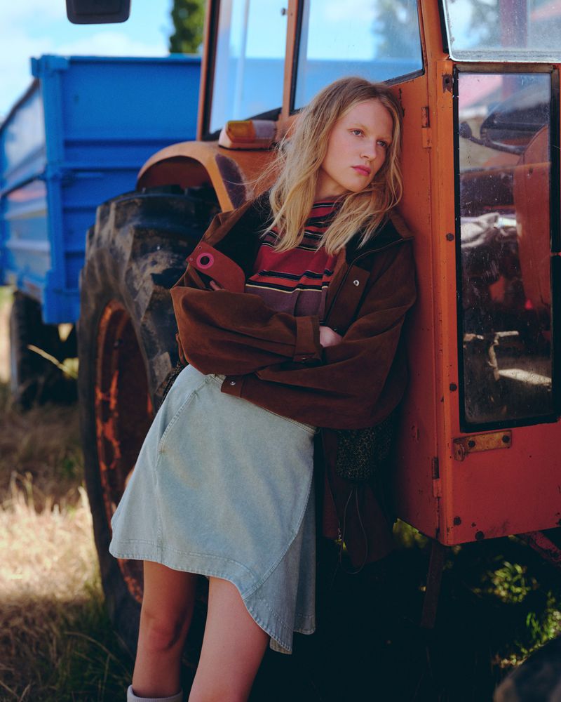 Woman leaning against a vintage tractor outdoors, wearing a brown jacket, striped shirt, and denim skirt in a rural setting.