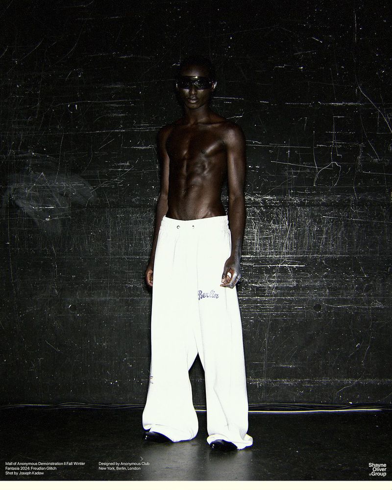 Shirtless male model with dark sunglasses poses in white pants against a dark wall, fashion, portrait, studio lighting, style.