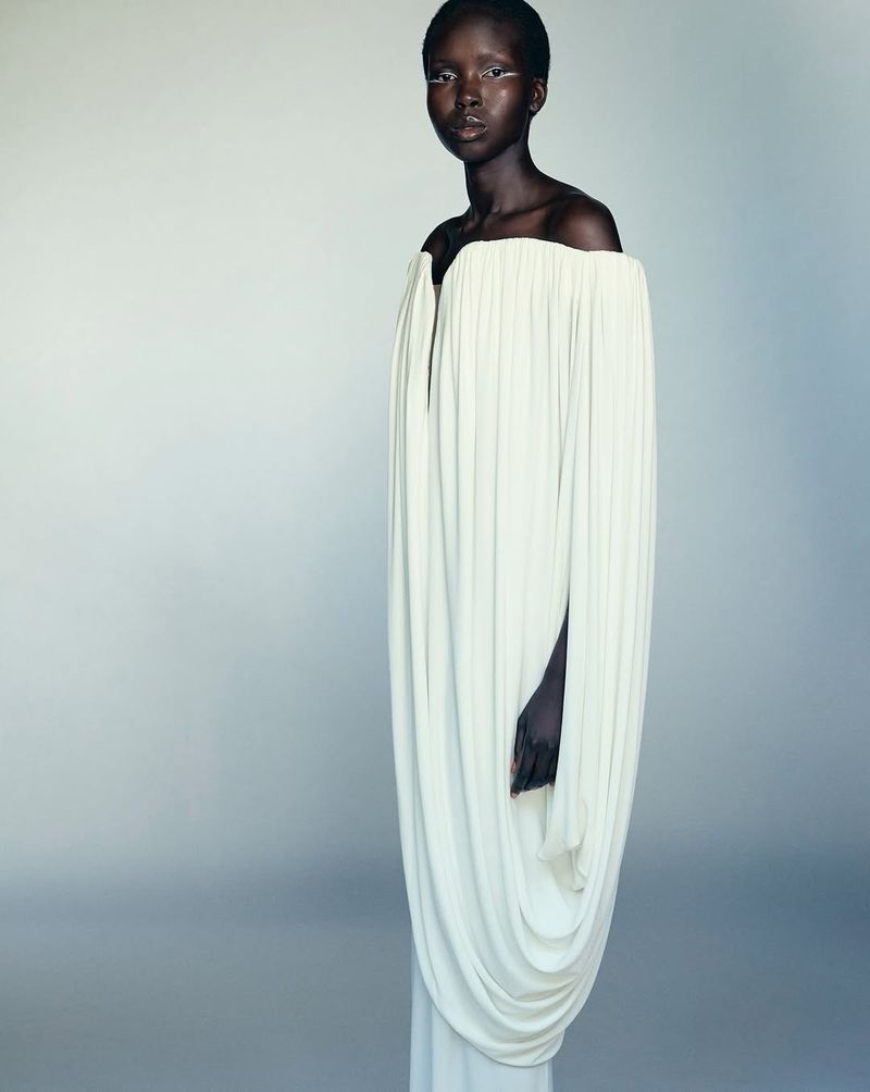 Woman with dark skin and a minimal white makeup posing in a flowing off-the-shoulder draped dress against a soft grey backdrop.