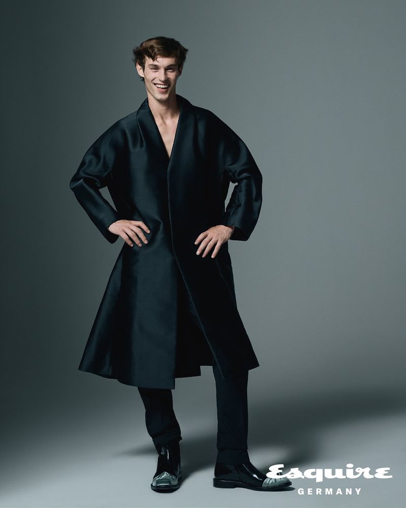 A full-length portrait of a smiling adult male wearing a black coat and pants. The man is set against a grey studio backdrop and has an easygoing pose.