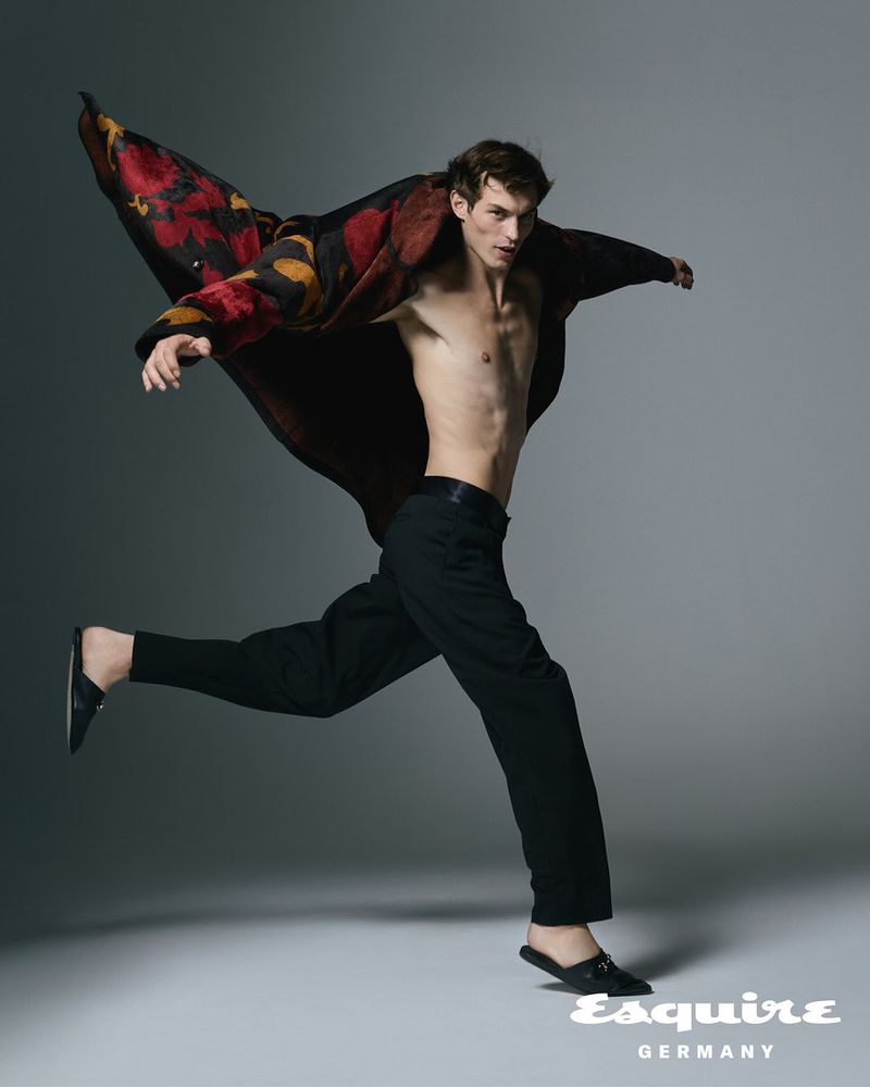 Male fashion model in motion, wearing a patterned coat, bare chest, and black pants against a neutral background for Esquire Germany.