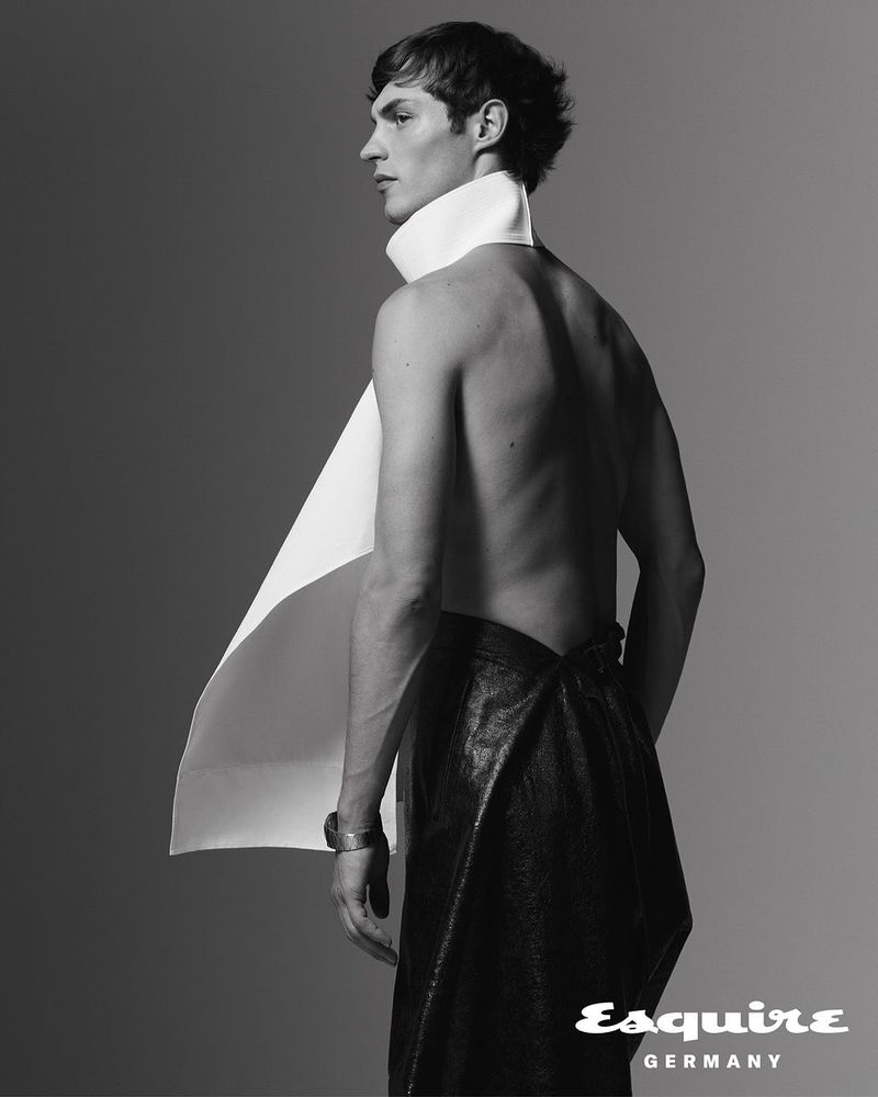 Monochrome portrait of a shirtless adult male with dark hair wearing a black skirt and white paper collar, posing in a photo studio.
