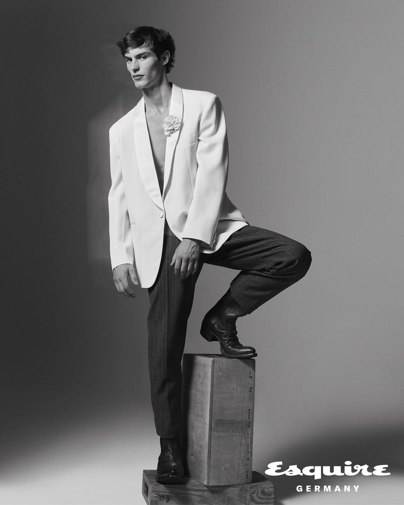 Black and white studio shot of a man in a suit jacket, pants, and boots posing on a wooden crate. Esquire GERMANY logo in the corner.
